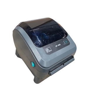 Peut inclure: Une imprimante d'étiquettes Zebra ZP 450 grise et noire. L'imprimante a un dessus noir, un bouton vert et une fente de sortie d'étiquettes. Le nom du modèle "ZP 450" est visible à l'avant de l'imprimante.
