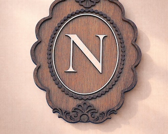 May include: A dark brown wooden wall plaque featuring an ornate, scalloped edge and a central oval frame. Inside the oval, a large, light-coloured capital letter N is displayed. The text Custom Initial • Handcrafted Wood appears below the plaque.