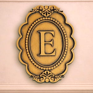 Op de afbeelding: Een goudkleurige wandplaat met een centraal ovaal frame met een kralenrand. De letter E staat prominent in het midden in een schreeflettertype. De plaat heeft een sierlijke, geschulpte rand met decoratieve krullen aan de boven- en onderkant.