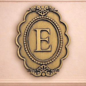 Può includere: Targa da parete decorativa color oro con la lettera "E" al centro. La targa ha una forma ovale con bordi smerlati ornati e decorazioni. La lettera "E" è centrata all'interno di una cornice ovale.