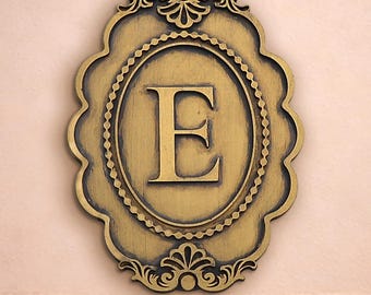 May include: A gold-coloured wall plaque with the letter "E" in the centre. The plaque has an oval shape with ornate, scalloped edges and decorative flourishes. The letter "E" is centered within an oval frame.