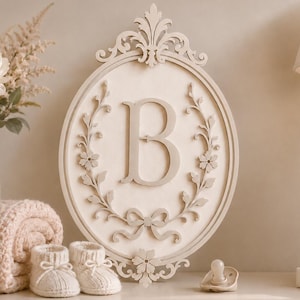Può includere: Una targa da parete di forma ovale color crema chiaro con una grande lettera maiuscola B al centro. La targa è decorata con un intricato bordo intagliato di viticci floreali e un fiocco sul fondo, sormontato da un fregio decorativo.