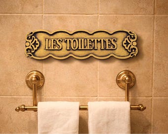 May include: A gold-coloured wooden sign with the French text "LES TOILETTES" above a brass towel rack with two white towels. The sign has a decorative, scalloped edge and ornate detailing. The sign is mounted on a tiled wall.