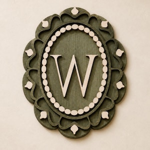 May include: An oval wall plaque featuring a dark olive green wooden frame with a decorative scalloped edge. The centre displays a large, light-coloured capital letter W surrounded by a border of small, circular light-coloured wooden beads.