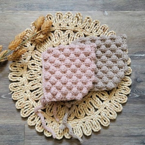 May include: Two crocheted baby hats, one pink and one brown, with a textured bubble stitch pattern. The hats are lying on a woven straw mat.