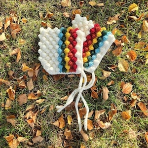 May include: A white crocheted hat with colorful bobbles in a rainbow pattern. The hat has a long white tie.