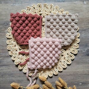 May include: Three crocheted baby bonnets in different colors: pink, beige, and burgundy. The bonnets are made with a textured stitch that creates a bumpy surface. The bonnets are arranged on a woven straw mat.