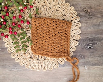 May include: A brown crocheted baby bonnet with a tie string. The bonnet is lying on a beige crocheted doily.