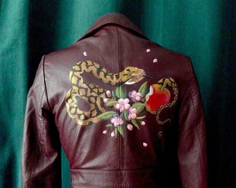 May include: A burgundy leather jacket with a painted design on the back. The design features a snake wrapped around a branch with pink flowers and a red apple.