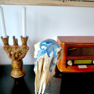 May include: A white animal skull with a blue and white painted design. The skull is on a black surface with a wooden radio and a candle holder in the background.