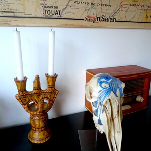 May include: A wooden candle holder with a decorative design and two white candles. The candle holder is on a black surface. A white animal skull with blue painted designs is on a black stand next to a wooden radio.
