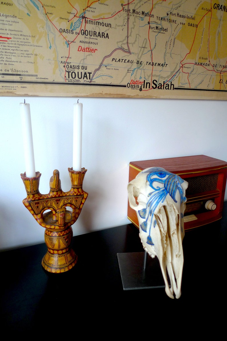May include: A wooden candle holder with a decorative design and two white candles. The candle holder is on a black surface. A white animal skull with blue painted designs is on a black stand next to a wooden radio.