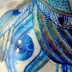 May include: Close-up of a white skull painted with intricate blue and gold designs. The skull features a large blue eye with gold accents and a detailed pattern of feathers and geometric shapes.
