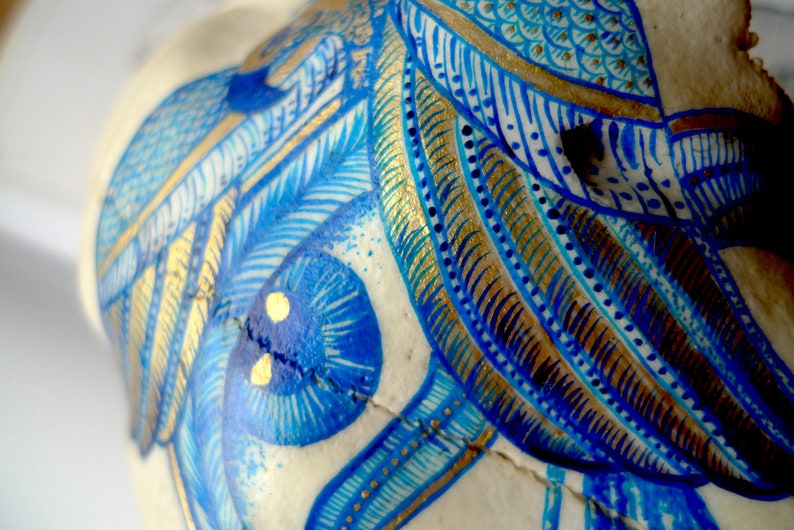May include: Close-up of a white skull painted with intricate blue and gold designs. The skull features a large blue eye with gold accents and a detailed pattern of feathers and geometric shapes.