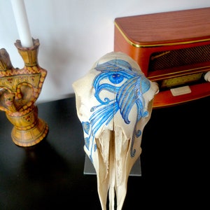 May include: A white animal skull with a blue and white painted design. The design features a large eye with intricate details and floral patterns.