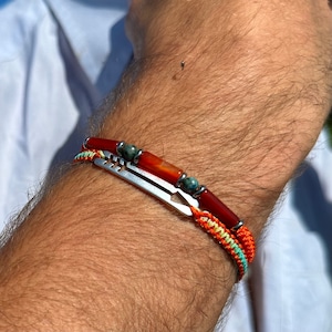 Puede incluir: Un brazalete con un dije de flecha de plata y cuentas rojas y verdes. El brazalete está en la muñeca de una persona.