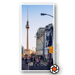 Könnte beinhalten: Ein Blick auf den Berliner Fernsehturm in der Ferne, mit einer Straßenszene im Vordergrund. Menschen gehen über einen Zebrastreifen, und es gibt ein Gebäude mit einem großen Wandbild eines Gesichts an der Seite.
