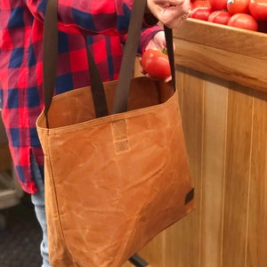 Puede incluir: Una bolsa de lona marr&oacute;n con asas de cuero marr&oacute;n. La bolsa la lleva una persona con una camisa de cuadros rojos y azules. La bolsa est&aacute; llena de tomates rojos.