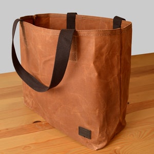 Puede incluir: Una bolsa de lona marr&oacute;n con correas negras. La bolsa est&aacute; sobre una superficie de madera.