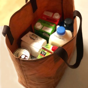 Puede incluir: Una bolsa de lona marr&oacute;n con asas negras llena de comestibles, incluyendo cartones de leche, cajas de jugo y latas.