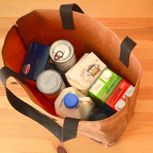 Puede incluir: Una bolsa de lona marr&oacute;n llena de comestibles, que incluyen una caja de pasta, dos latas de sopa, una caja de leche, una caja de az&uacute;car y un recipiente de caldo de verduras.