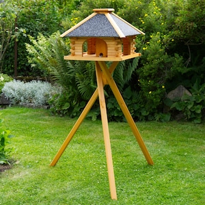 Op de afbeelding: Een houten vogelvoederhuisje met een grijs dak van shingles en een driehoekige basis. De voederbak heeft twee voederplaatsen en is gemonteerd op een hoge houten driepoot.