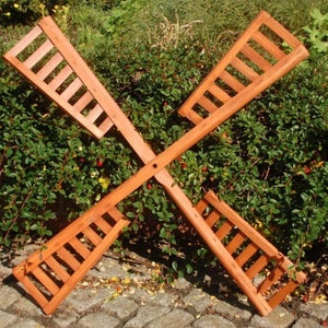 Könnte beinhalten: Eine hölzerne Windmühle mit vier Flügeln, die jeweils ein Lamellendesign aufweisen. Die Windmühle ist braun und liegt auf einem Kiesbett.