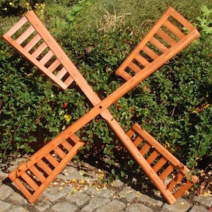 Könnte beinhalten: Eine Windmühle aus Holz mit vier Flügeln, die jeweils ein Lamellendesign aufweisen. Die Windmühle ist braun und liegt auf einem Kiesbett.