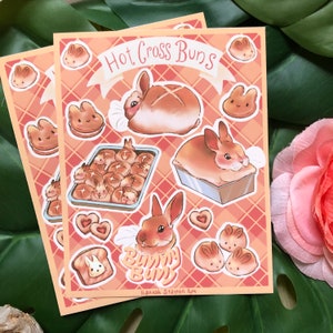 Könnte beinhalten: Zwei Kunstdrucke mit Illustrationen von Hot Cross Buns und hasenförmigem Gebäck. Die Drucke haben einen pfirsichfarbenen und roten Karohintergrund. Der Text "Hot Cross Buns" steht auf einem Banner, und "Bunny Buns" steht ebenfalls auf dem Druck.