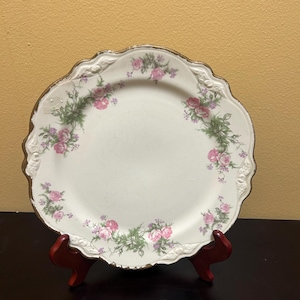 Può includere: Un piatto in porcellana bianca con un bordo ondulato e un bordo dorato. Il piatto è decorato con un motivo floreale rosa e verde.