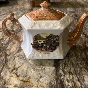 May include: White and brown ceramic teapot with a detailed handle and spout. The lid and handle feature a brown patterned design. The teapot has a rectangular shape with a scenic image and the text "Old Coach House, Bristol".