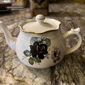 May include: Cream-colored ceramic teapot with a lid, featuring a black rose floral design. The teapot has a gold trim around the lid and spout. The handle is curved, and the teapot sits on a marble-like surface.