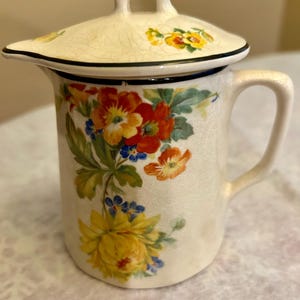 Puede incluir: Una jarra de crema de cerámica blanca con un diseño floral. La jarra tiene una tapa con asa y un pico. El diseño floral presenta flores amarillas, rojas, naranjas y azules.