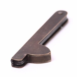 Könnte beinhalten: Ein Multitool aus Messing mit einem Flaschenöffner und einer Klinge. Das Werkzeug ist zusammengeklappt und hat eine abgenutzte, gealterte Optik.