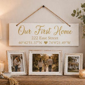 May include: A cream-coloured wooden sign with gold lettering reading "Our First Home", the address "222 East Street", and coordinates. Below are three framed photos with white distressed frames. A lamp and candle are on the left.