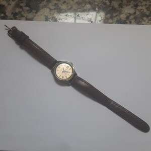 K&ouml;nnte beinhalten: Eine braune Lederarmbanduhr mit goldenem Zifferblatt und r&ouml;mischen Ziffern als Stundenmarkierungen.
