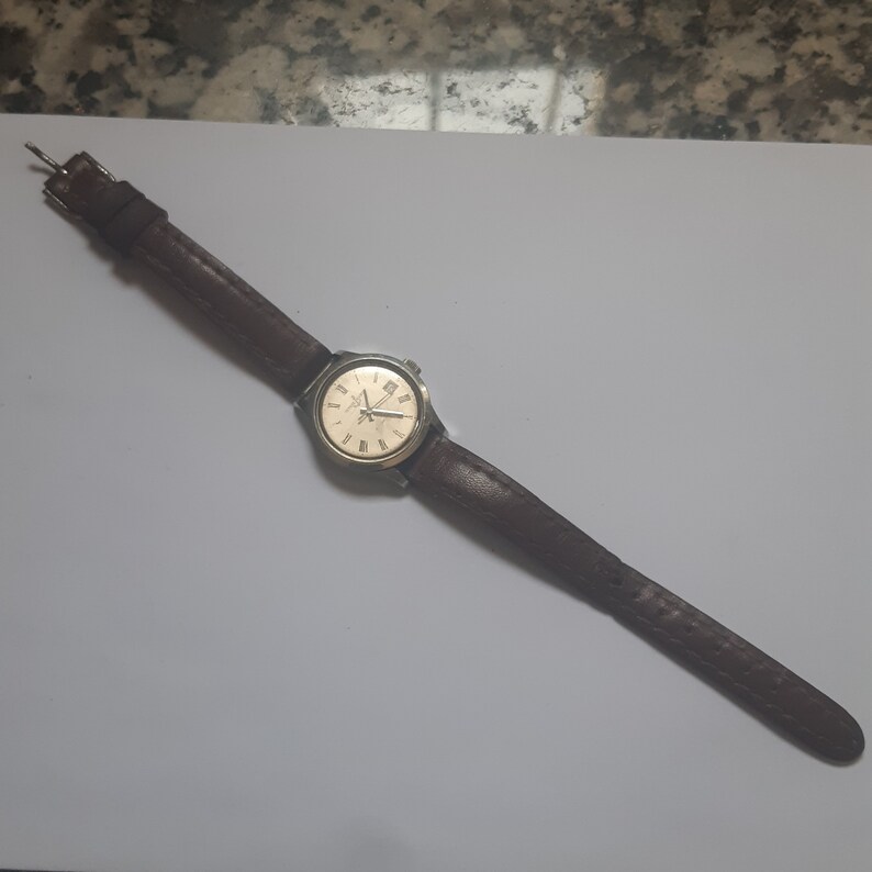 K&ouml;nnte beinhalten: Eine braune Lederarmbanduhr mit goldenem Zifferblatt und r&ouml;mischen Ziffern als Stundenmarkierungen.