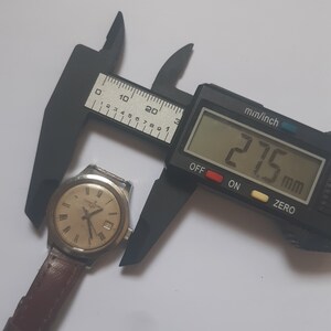 K&ouml;nnte beinhalten: Eine silberne Armbanduhr mit wei&szlig;em Zifferblatt und schwarzen r&ouml;mischen Ziffern als Stundenmarkierungen. Die Uhr wird mit einer digitalen Schieblehre gemessen, die 27,5 mm anzeigt.