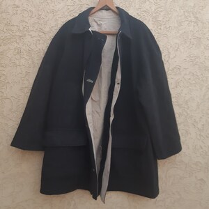 May include: A dark navy blue coat with a light beige interior hangs on a wooden hanger. The coat has a classic design with a collar, buttons, and a long length. The coat's exterior is a solid, dark color, while the interior lining is a lighter shade.