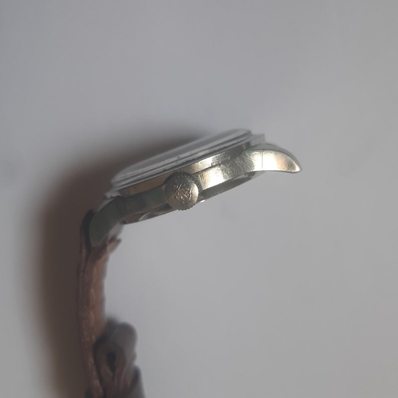 K&ouml;nnte beinhalten: Eine silberne Armbanduhr mit einem braunen Lederarmband. Die Uhr hat ein rundes Zifferblatt und eine silberne Krone.