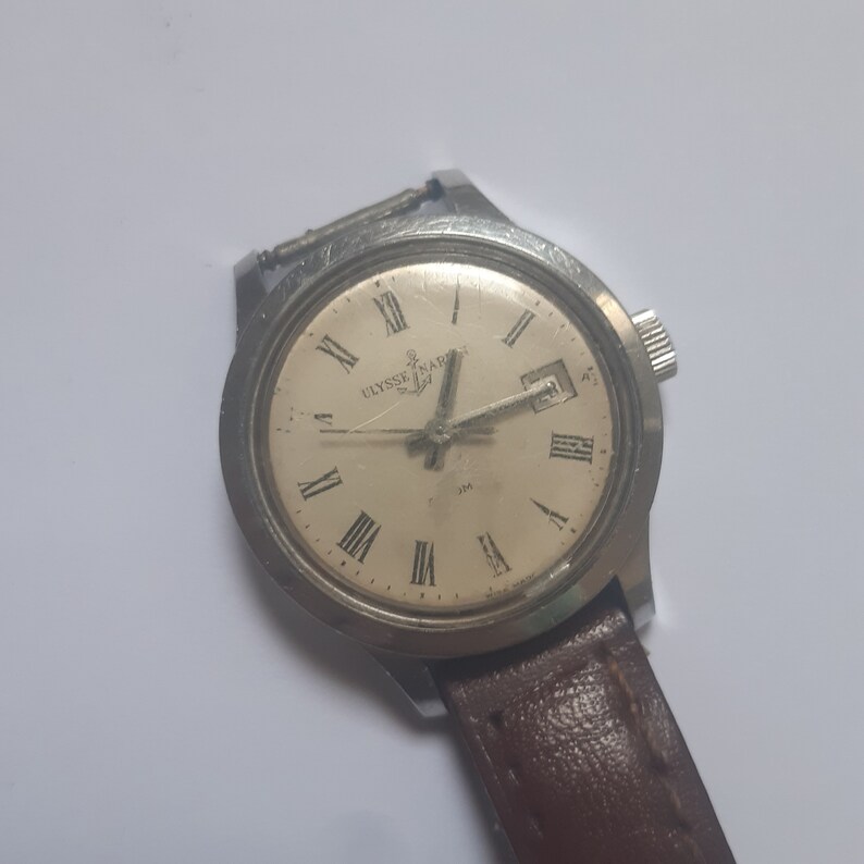 K&ouml;nnte beinhalten: Eine silberne Armbanduhr mit wei&szlig;em Zifferblatt und schwarzen r&ouml;mischen Ziffern als Stundenmarkierungen. Die Uhr hat ein braunes Lederarmband und tr&auml;gt die Aufschrift "Ulysse Nardin" auf dem Zifferblatt.