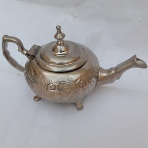 May include: A silver teapot with an ornate design and a lid. The teapot has three legs and a spout.
