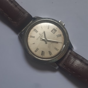 K&ouml;nnte beinhalten: Eine silberne Armbanduhr mit wei&szlig;em Zifferblatt und r&ouml;mischen Ziffern als Stundenmarkierungen. Die Uhr hat ein braunes Lederarmband und ein Datumsfenster bei 3 Uhr. Das Zifferblatt der Uhr zeigt "Ulysse Nardin".