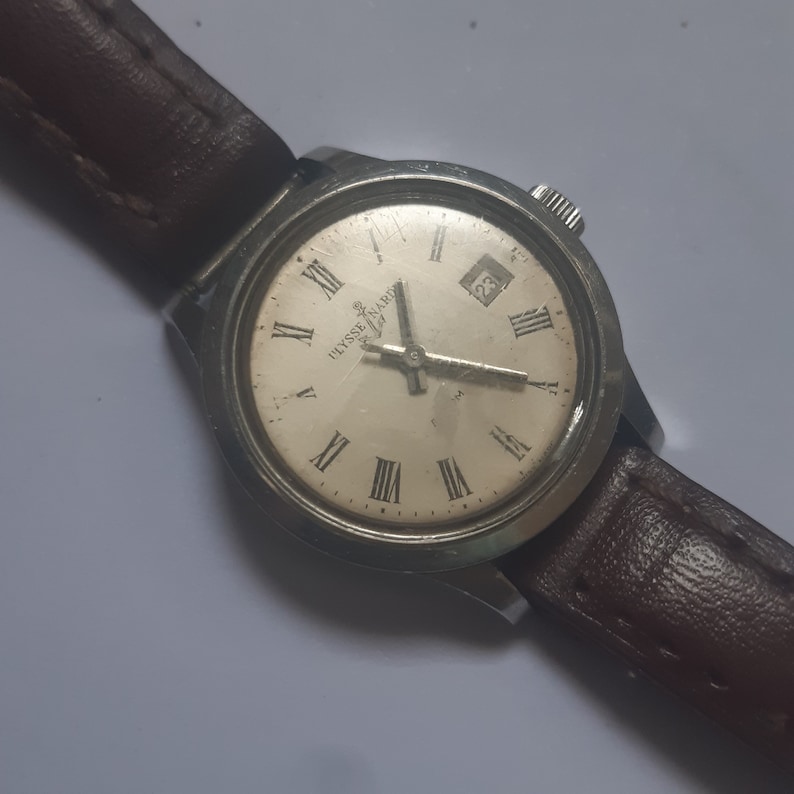 K&ouml;nnte beinhalten: Eine silberne Armbanduhr mit wei&szlig;em Zifferblatt und r&ouml;mischen Ziffern als Stundenmarkierungen. Die Uhr hat ein braunes Lederarmband und ein Datumsfenster bei 3 Uhr. Das Zifferblatt der Uhr zeigt "Ulysse Nardin".
