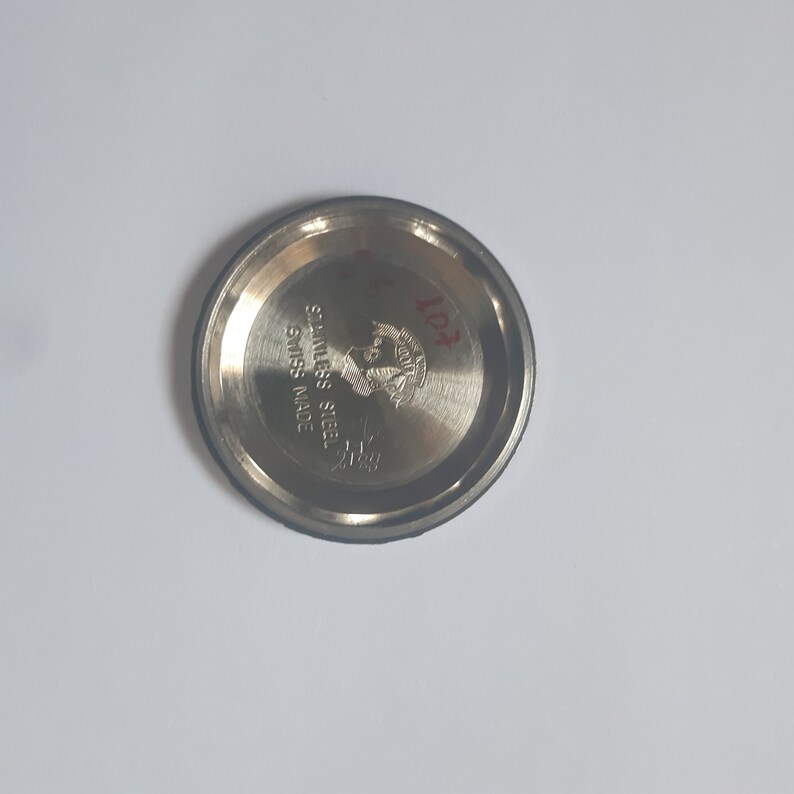 K&ouml;nnte beinhalten: Nahaufnahme eines silbernen Uhrenbodens mit der Gravur "STAINLESS STEEL" und "SWISS MADE". Der Boden hat auch ein Logo mit dem Text "11PR".