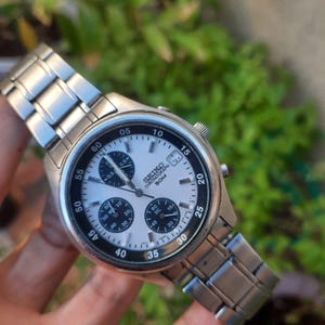 May include: A silver Seiko chronograph wristwatch with a white face and black markings. The watch has a date window at the 3 o'clock position and a tachymeter scale around the outer edge of the face. The watch is on a silver metal band.