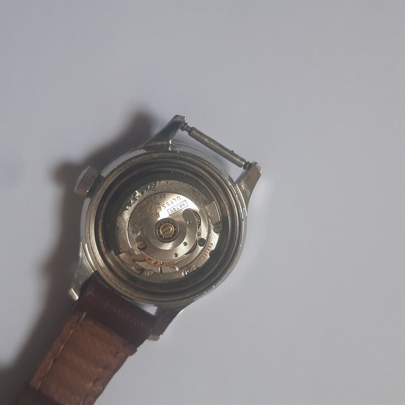 K&ouml;nnte beinhalten: Die R&uuml;ckseite einer silbernen Armbanduhr mit einem braunen Lederarmband. Das Zifferblatt der Uhr ist offen und zeigt das Innenleben der Uhr. Der Text "USSATO" und "17 Jewels" ist auf dem Zifferblatt der Uhr sichtbar.