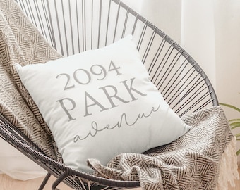 Puede incluir: Una silla de alambre negra con un cojín blanco que dice "2094 PARK avenue" en letras grises. Una manta estampada beige y blanca con flecos está sobre la silla. Una planta en maceta está en el fondo.