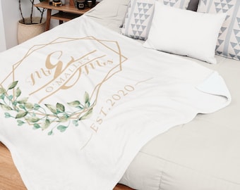 May include: A white blanket with a gold geometric design and the text "Mr & Mrs O'Malley EST. 2020" is displayed on a bed. The blanket is decorated with green leaves. Decorative pillows and a framed art piece are also visible.
