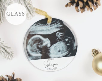 May include: A white round glass ornament with a black and white ultrasound image of a baby. The ornament has a gold ribbon and the text "BABY Johnson JUNE 2022" printed on it.